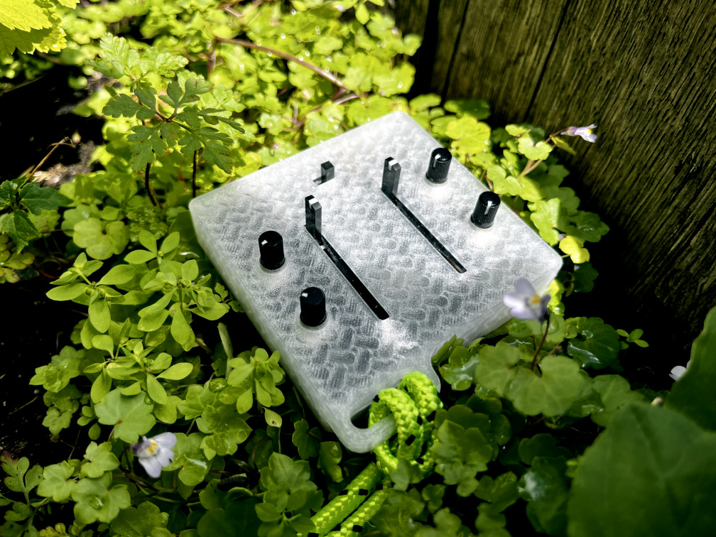 A photo of Lore, a small semi-transparent synthesizer made of bioplastic, sitting on top of some small green plants next to a wooden planter box